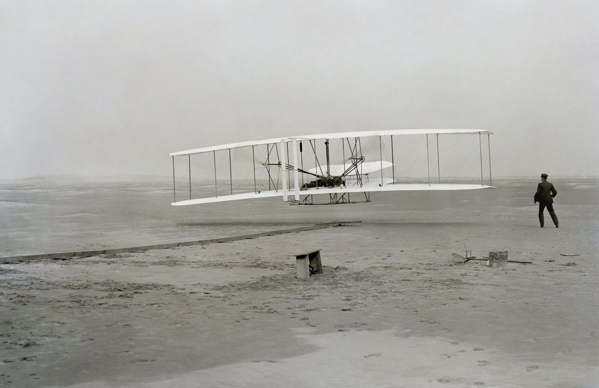 The Wright Flyer a few feet off the ground on its first flight at Kitty Hawk in 1903, Wilbur Wright running alongside on the sand